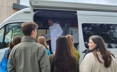 A scientist in a white coat inside a mobile laboratory talking to a group of young people