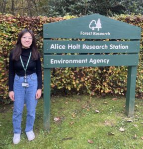 A young woman standing next to a sign reading 'Alice Holt Research Station'