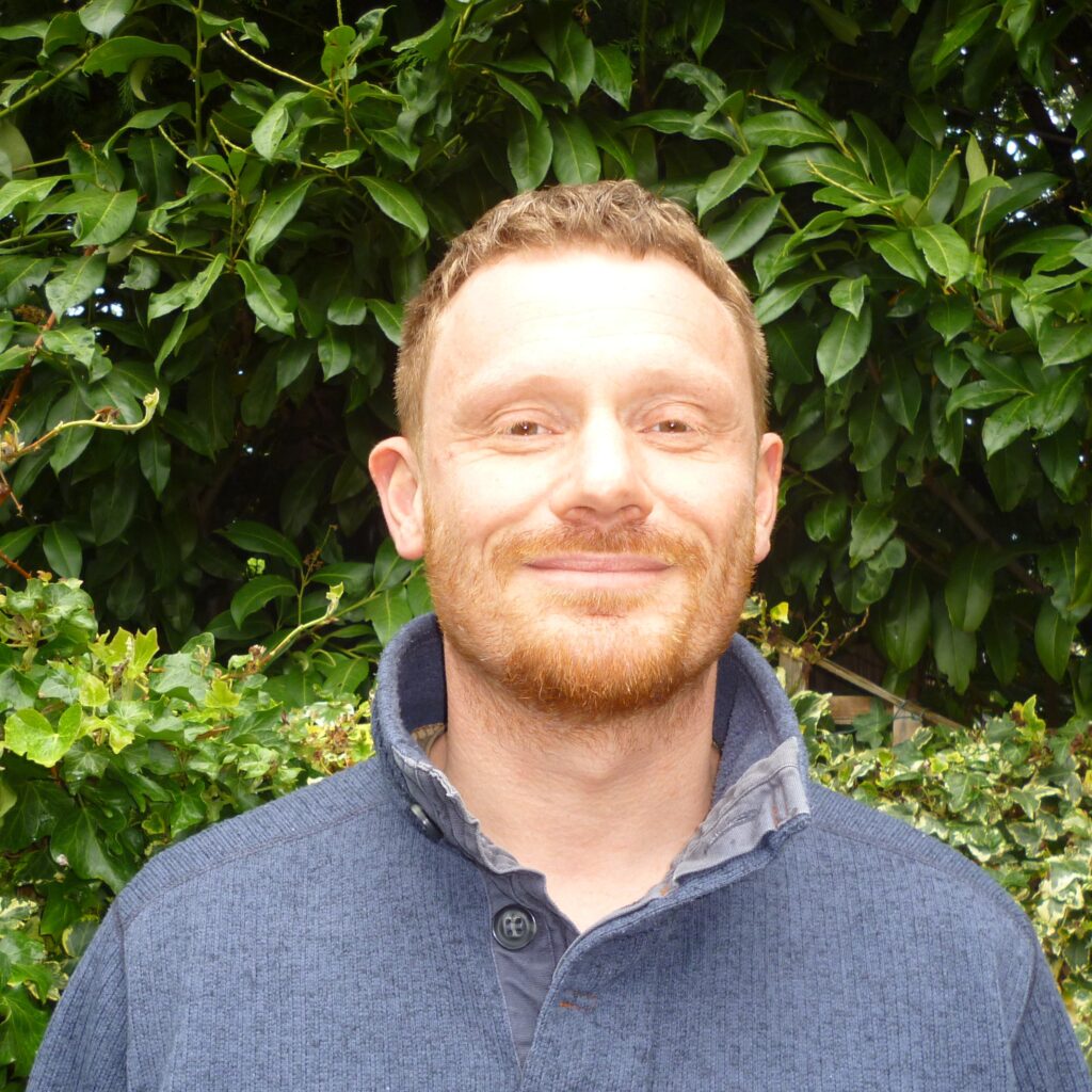 Image of a man with ginger hair and beard, wearing a dark blue open collar fleece, against a backdrop of dark green bushes