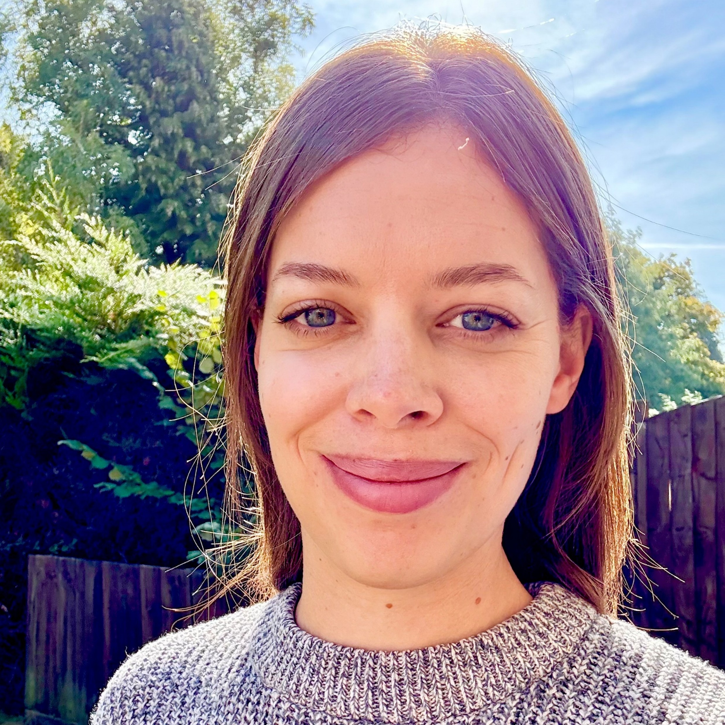 Jess Allan A smiling woman with long brown hair, wearing a grey woolly jumper and is standing in front of a fence, grass and some bushes against a sunny blue sky.