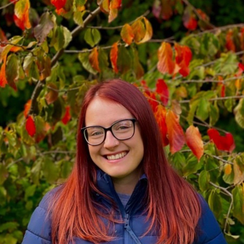 Milly - Photo A woman with long, dark red hair, and black glasses, wearing a dark blue jacket, smiling against a background of a tree covered in colourful leaves,