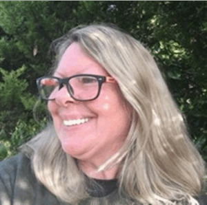 A smiling woman with long blonde hair, and black glasses. She is facing towards the left, and is wearing a dark grey-brown jumper on top of a black tshirt. She is stood against an out of focus background of bushes.
