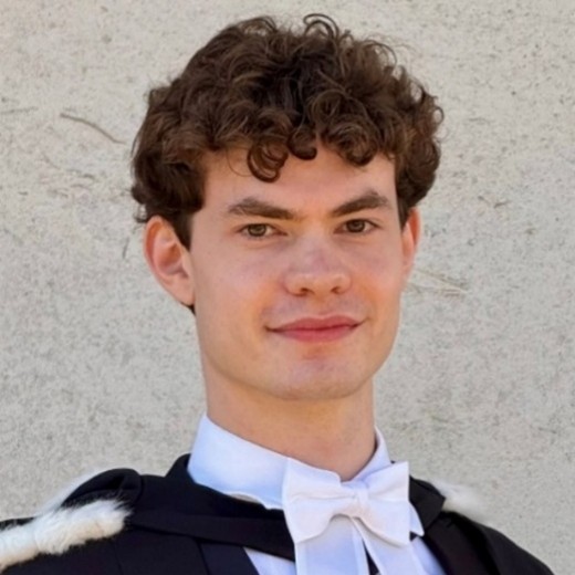 A smiling woman with long blonde hair, and black glasses. She is facing towards the left, and is wearing a dark grey-brown jumper on top of a black tshirt. She is stood against an out of focus background of bushes.
