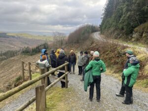 A group of 13 people dressed in winter clothing (thick jackets, walking trousers and boots), stood on a path to the left of a forest, with a valley in the background.