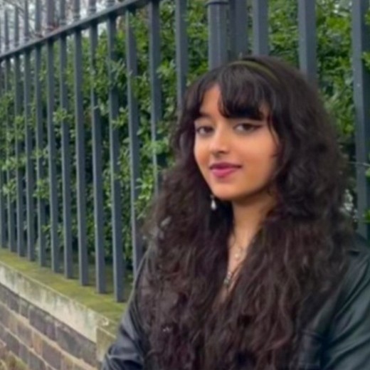 A woman with long, dark red hair, and black glasses, wearing a dark blue jacket, smiling against a background of a tree covered in colourful leaves,