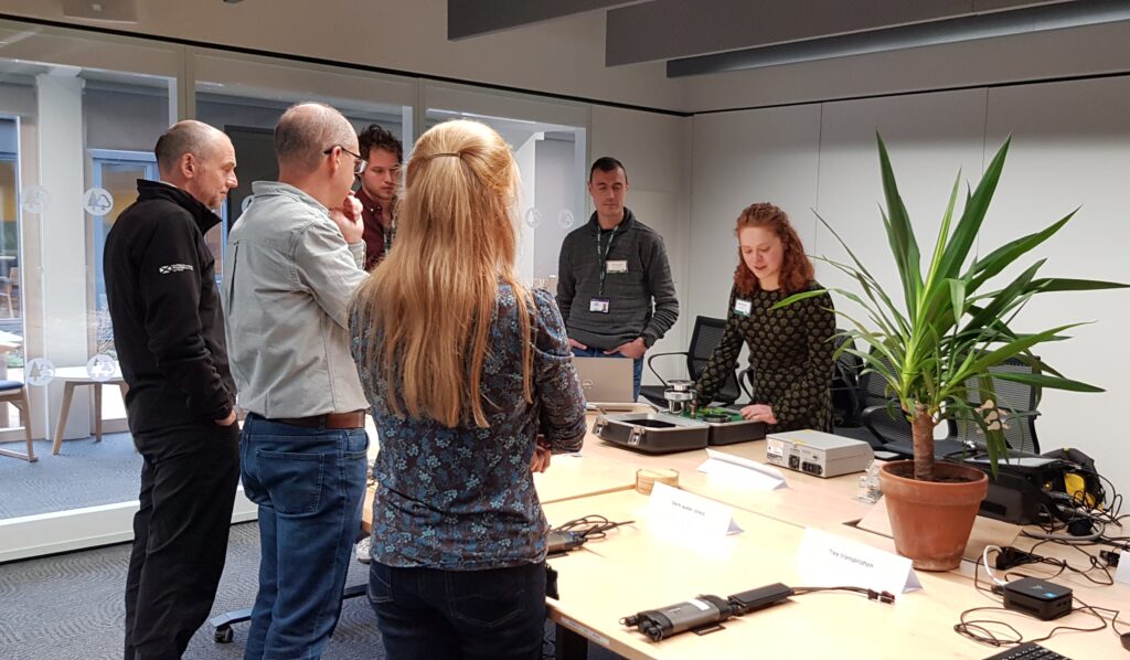 A person with wavy red hair in a black dress leading a demonstration using scientific equipment to assess tree stem and leaf water stress, surrounded by five other people.