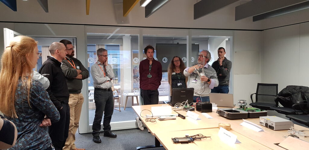 A person with grey hair wearing glasses, a grey jumper, and blue jeans holding up a piece of equipment used to assess wind speed and direction. Eight other people are in the room watching the demonstration.