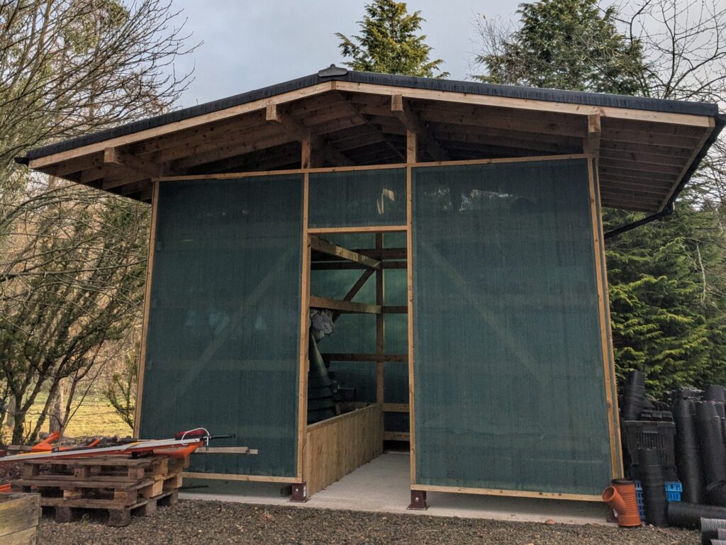 A small wooden building with a roof which extends further out to the left and right than the walls. The walls are made from a thin translucent blue/grey material. Inside there are wooden beams with small white material bags hanging off of them. In the foreground there are three wooden pallets with some other wood and plastic materials to the left of the building, and some black plastic pots for planting to the right of the building.
