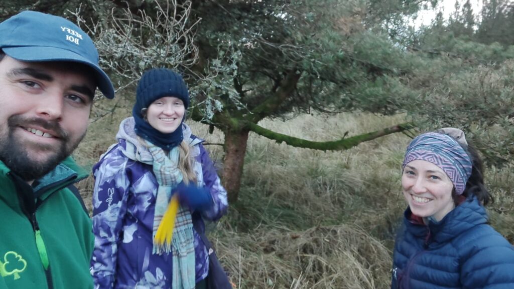 Three people stood against a background of a tree, in a field with long grass. The person on the left is taking the selfie of the three people. They have a dark beard and moustache and is wearing a blue cap and a green zip up fleece. The person in the middle has long blonde hair and is wearing a light and dark blue flower patterned winter jacket. They are also wearing a grey and brown patterned scarf, and a navy hat and snood. The person on the right has dark brown hair tied back in a pony tail, and is wearing a multi-coloured patterned headband, and a navy puffer jacket. All three are smiling at the camera in a selfie pose.