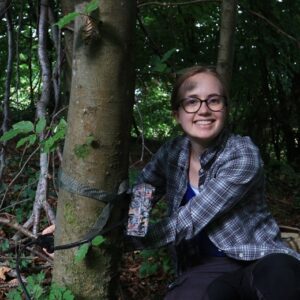 A person sitting in a wooded area with one arm secured to a tree using a strap. The individual is wearing an outdoor jacket and sitting on the ground among leaves and branches.
