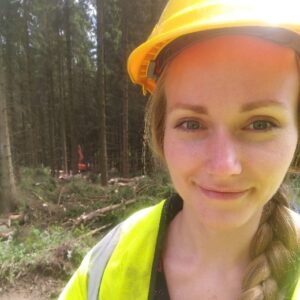 A person with long blonde hair in a plait, wearing a yellow hard hat and a high‑visibility jacket stands in a forested area holding a metal water bottle. Tall trees, cut branches, and forestry equipment are visible in the background.