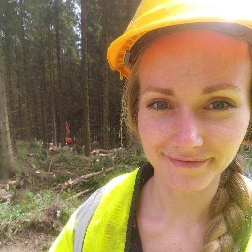 A person with long blonde hair in a plait, wearing a yellow hard hat and a high‑visibility jacket stands in a forested area holding a metal water bottle. Tall trees, cut branches, and forestry equipment are visible in the background.