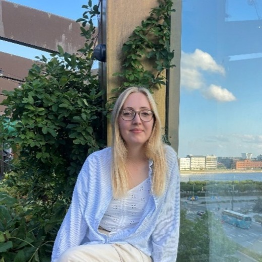 A person with long blonde hair sitting outdoors beside a wooden post covered in climbing plants. They are wearing a light blue open shirt over a white eyelet-style top and light-coloured trousers. Behind them, there is a view of a cityscape with buildings, roads, and buses near a body of water under a blue sky with a few clouds.