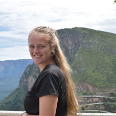 A person with long, blonde hair stands outdoors in front of a mountainous landscape. The sky is partly cloudy, and green cliffs and ridges stretch into the distance behind them.