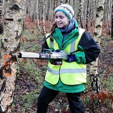 A person wearing a bright yellow safety vest, winter clothing, and gloves stands in a birch woodland while using a handheld tool against the trunk of a tree.