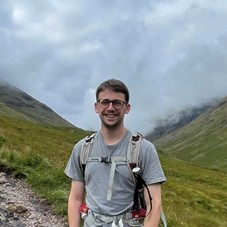 A person wearing a grey T-shirt and hiking backpack stands on a path surrounded by grass covered mountains, with misty clouds hanging low over the landscape.