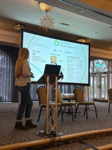 A person stands beside a lectern and large presentation screen in a conference room. The screen displays a slide titled “Centre for Forest Protection,” featuring logos, text sections, and circular graphic elements. Several chairs are arranged on the stage behind the speaker, and large windows in the background let in natural light.