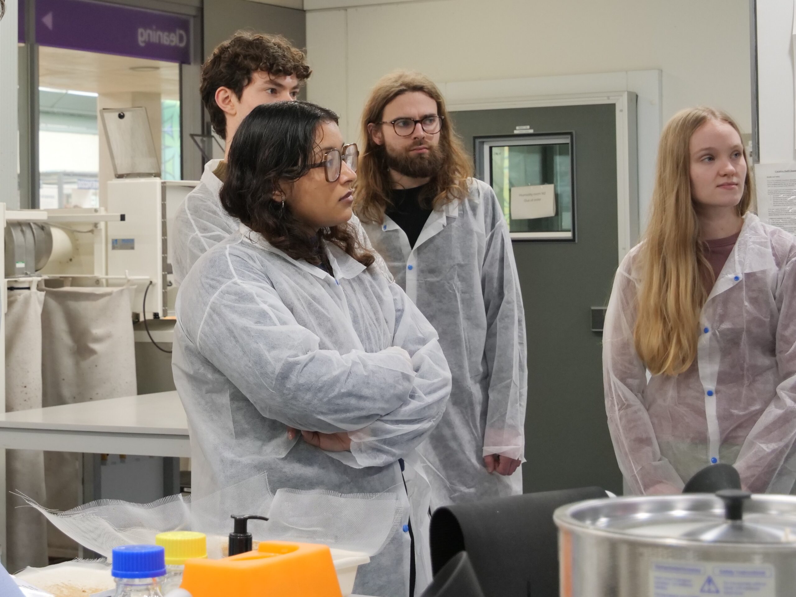 A group of people wearing lab coats stand together in a laboratory, appearing to listen to someone outside the frame. The room contains lab benches, equipment, and bright overhead lighting. Various containers and supplies are visible on the work surface in the foreground.
