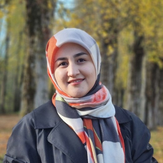 A person wearing an orange, black, and white, patterned headscarf and a dark coat standing outdoors on a tree-lined path during autumn, with tall trees displaying yellow and golden leaves in the background.