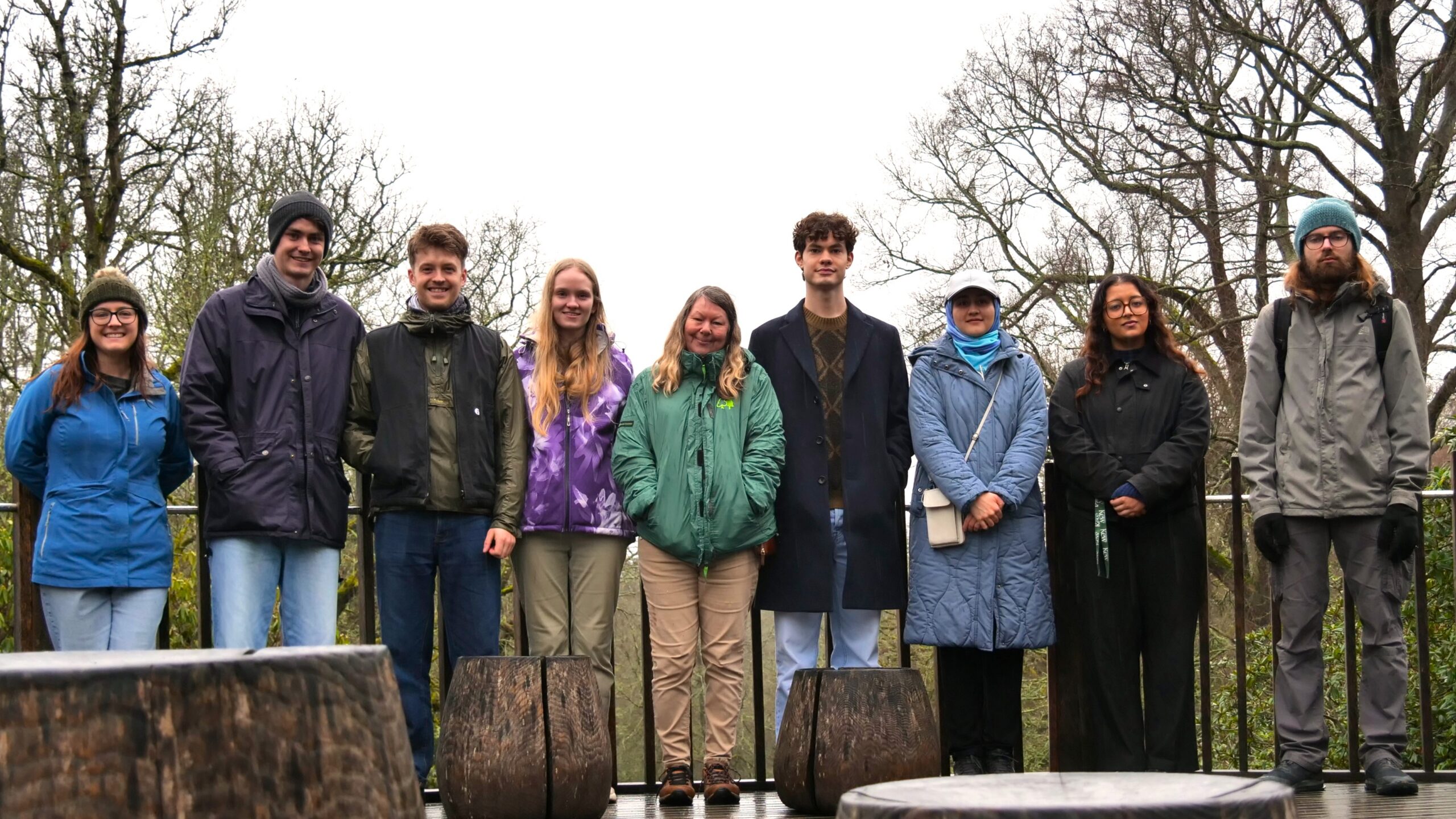 A group of nine people standing in a row outdoors on an overcast day. They are dressed in winter jackets, coats, hats, and boots, standing on a wooden platform with large tree stumps in the foreground. Leafless trees fill the background, indicating a cold season.