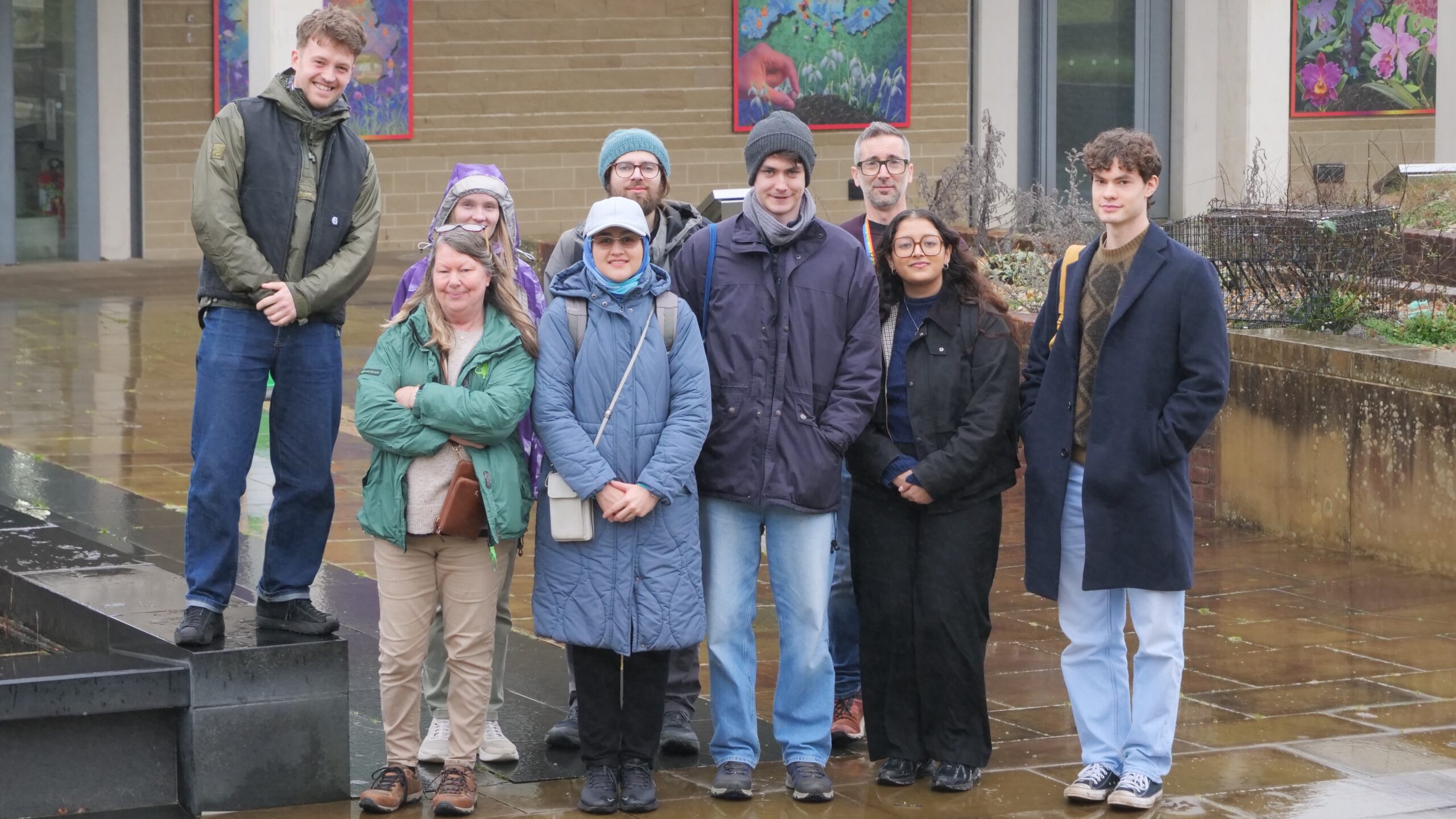 A group of nine people stand outdoors on a wet, paved area in front of a building with colourful artwork in the windows. They are dressed in winter clothing, including coats, hats, and scarves, and they smile for the camera. The scene is overcast and rainy.