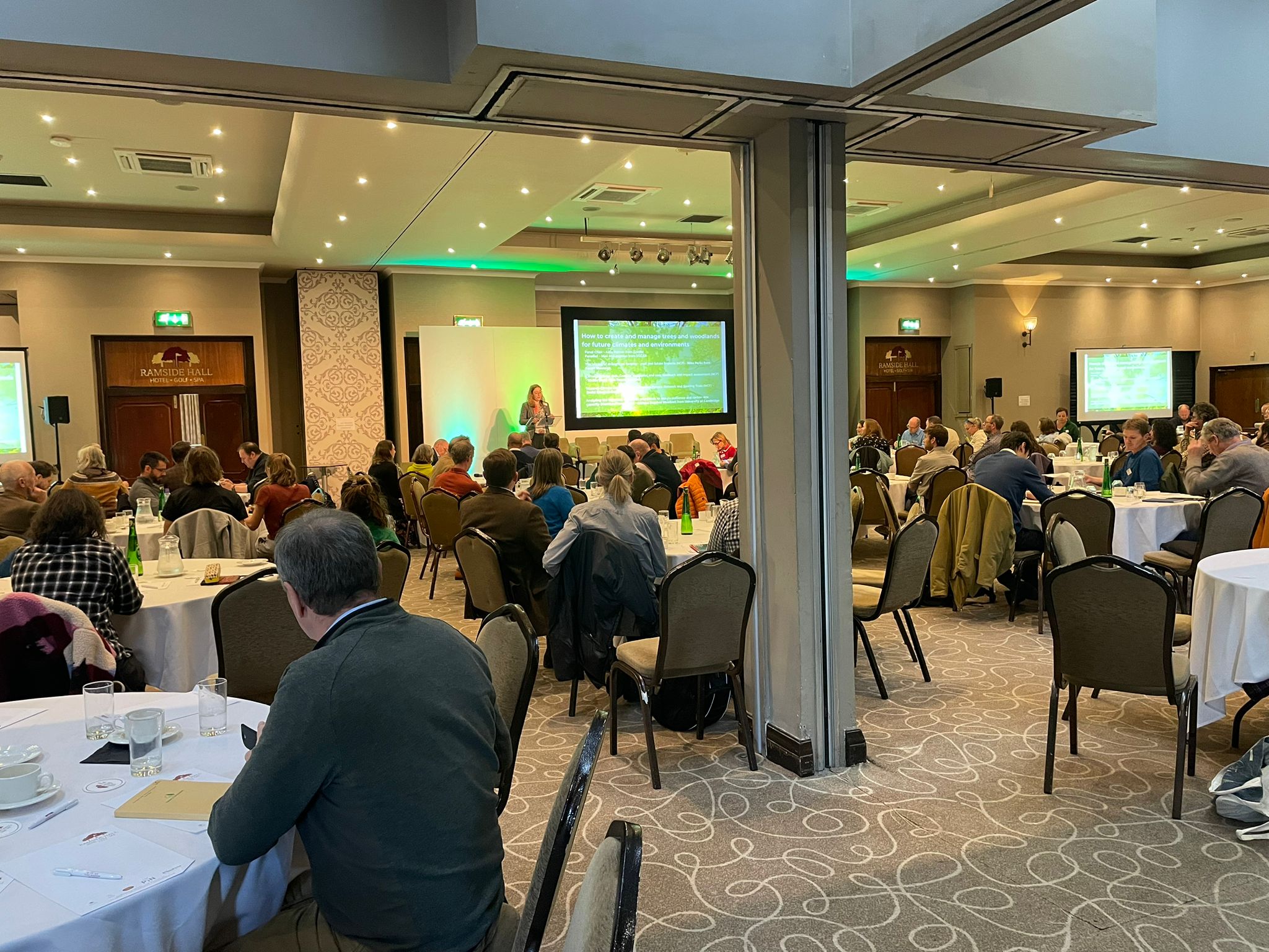 Britain’s Forest Future © Deborah Talbot, Future of UK Treescapes A large conference room with round tables where attendees are seated and listening to a speaker at the front. Two projection screens display slides with green and yellow graphics. The room is well‑lit with ceiling spotlights and neutral décor. People are engaged in discussion and facing the stage.