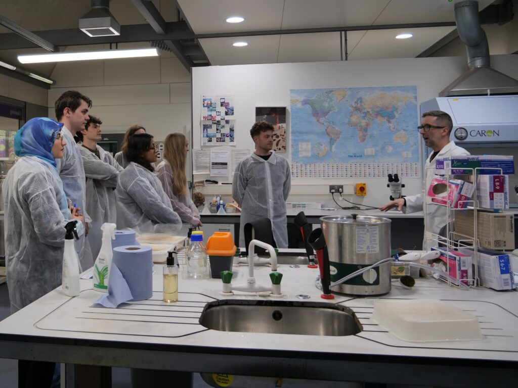A group of people wearing disposable lab coats stand together in a laboratory, listening to an instructor who is speaking and gesturing toward equipment. The room contains sinks, bottles, tools, and scientific apparatus on the central workbench. A large world map and various notices are displayed on the wall behind them, and boxed lab supplies are stacked on shelves to the right.