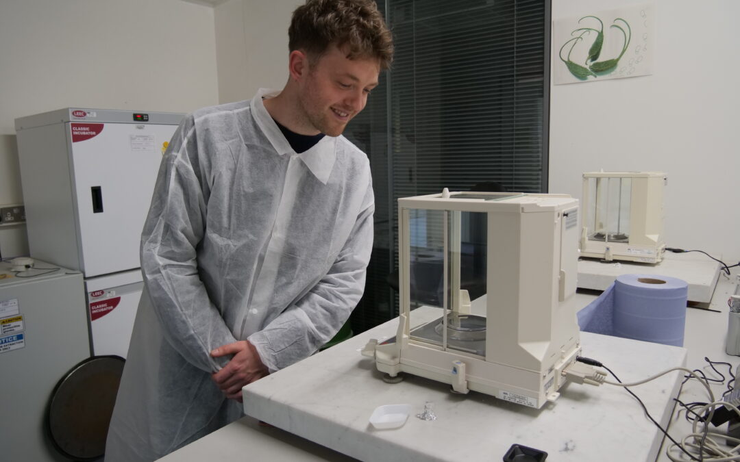A person wearing a white lab coat stands at a laboratory workbench, looking down at a digital precision scale enclosed in a transparent case. Small objects and tools lie on the counter nearby, with scientific equipment, including an incubator and cables, visible in the background of the lab.