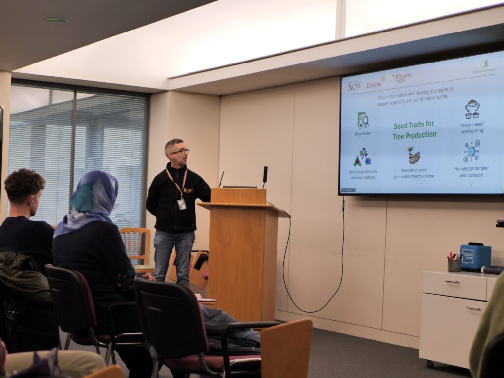 A person stands at a wooden podium giving a presentation to an audience seated in a conference room. A large screen displays a slide titled “Seed Traits for Tree Production,” featuring icons and brief text about seed traits and conservation. The audience members face the presenter, and the room has large windows and neutral-coloured walls.