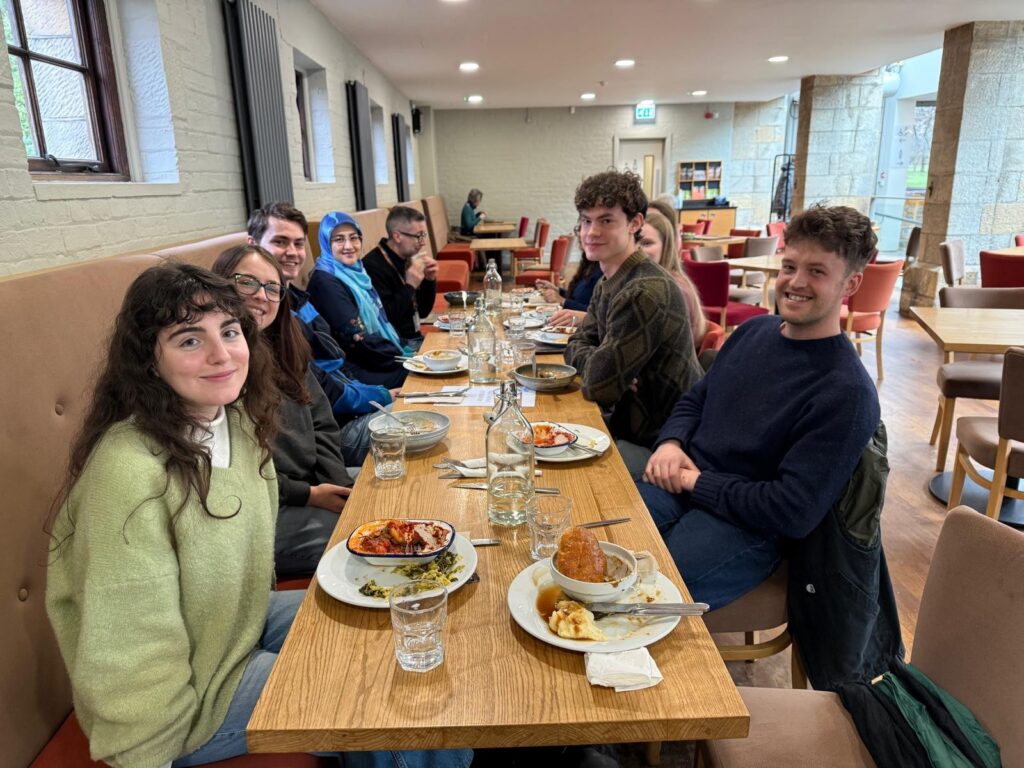 A group of people sit around a long wooden table in a café or dining hall, finishing a meal together. Plates with leftovers, glasses, and water bottles are scattered across the table. The setting has light-coloured brick walls, wooden floors, and cushioned seating, with large windows bringing in natural light. The group appears relaxed and engaged in conversation.