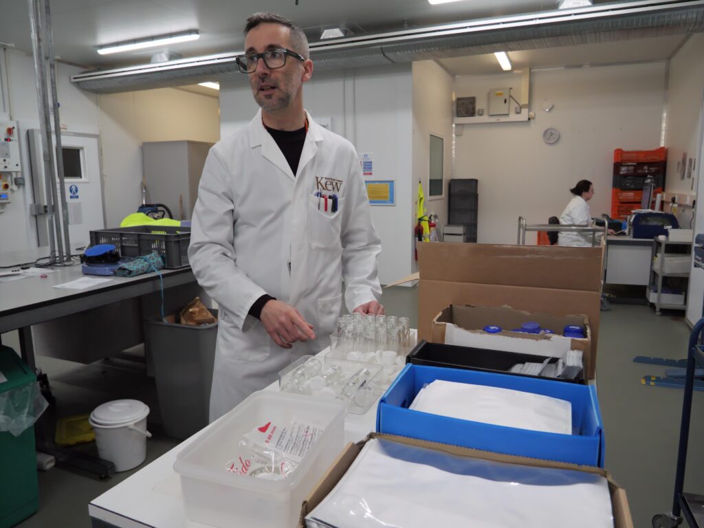 A person wearing a white lab coat stands at a workstation in a laboratory, arranging glass vials on a counter. Boxes containing lab materials and plastic containers are on the table in front of them. The room has industrial lighting, lab equipment, and another person working in the background.