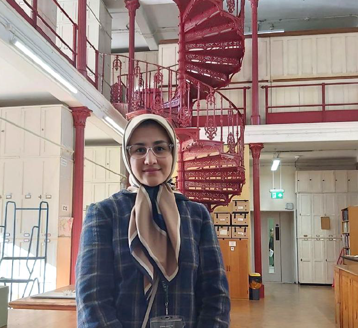 A person wearing glasses, a light-coloured head covering, and a dark plaid jacket stands indoors in front of an ornate red metal spiral staircase. The space appears to be a large institutional interior with cream-coloured walls, wooden floors, railings, storage cabinets, and shelves in the background. The staircase rises behind the person across two levels, with decorative railings and columns. An exit sign and stacked boxes are visible further back in the room.