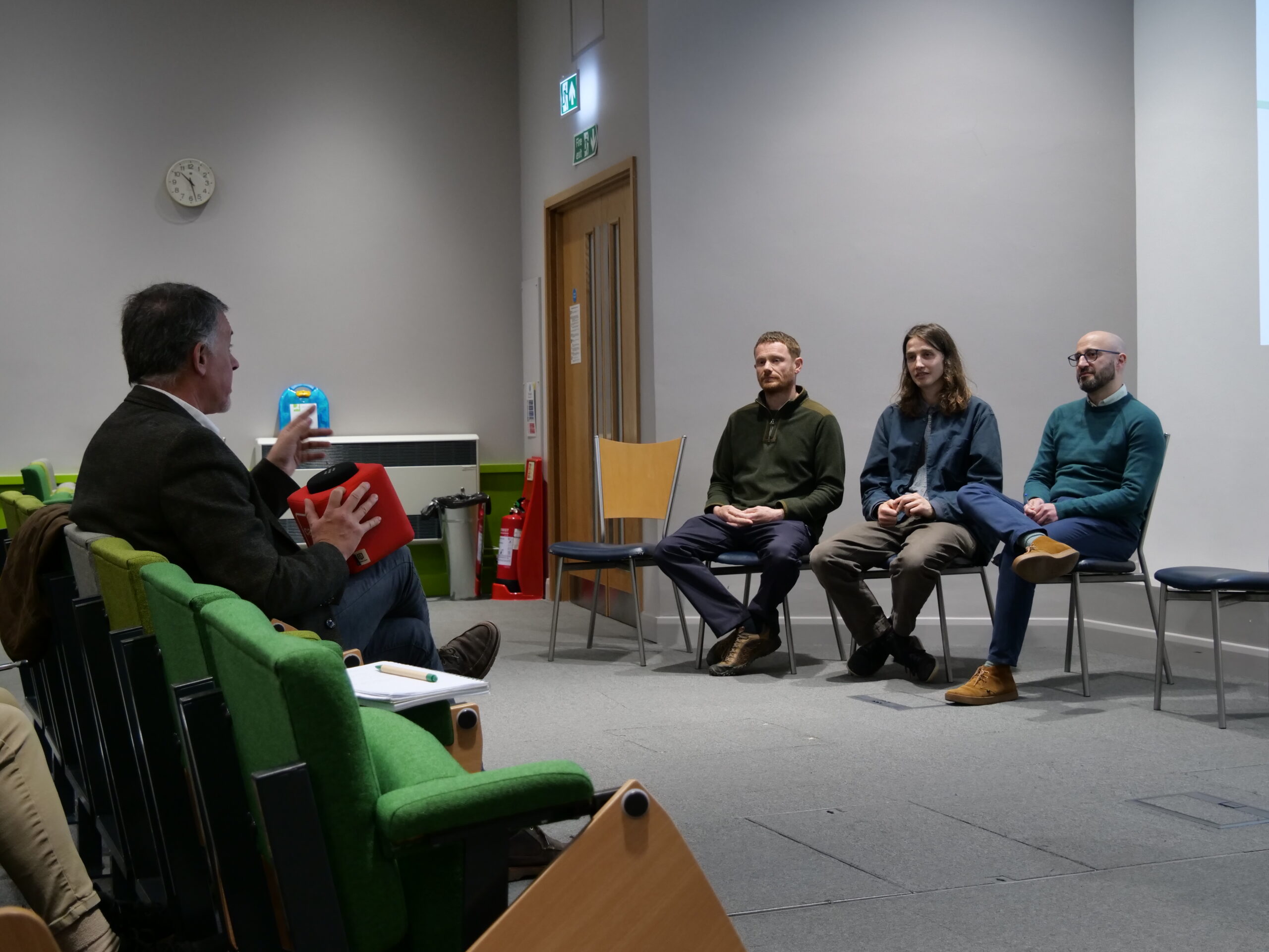 A small panel discussion taking place in a lecture room. Three people sit on simple chairs arranged in a row at the front of the room, facing an audience. Another person sits to the left in a green auditorium chair, holding a red ‘Catch box’ microphone and gesturing while speaking toward the panel. The room has grey walls, a wall clock, an exit sign above a wooden door, and safety equipment near the wall. The setting suggests a question‑and‑answer session or facilitated discussion.