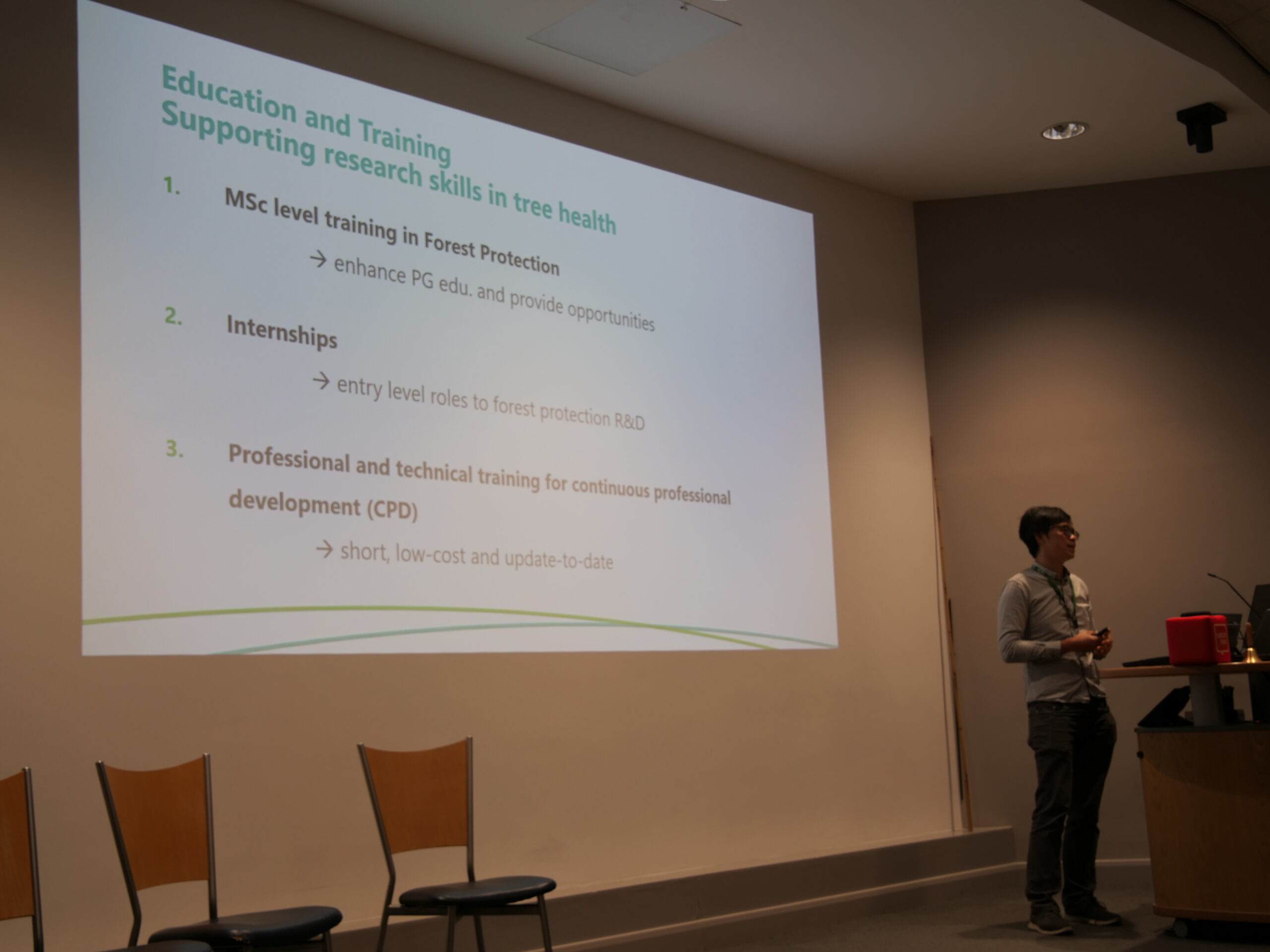 A large projection screen at the front of a lecture room displays a slide titled ‘Education and Training: Supporting research skills in tree health,’ with a numbered list including MSc‑level training in forest protection, internships, and professional and technical training for continuing professional development. A person stands to the right of the screen near a lectern with a laptop and microphone, facing the projected slide. Several empty chairs are visible in the foreground, and ceiling lights illuminate the room.