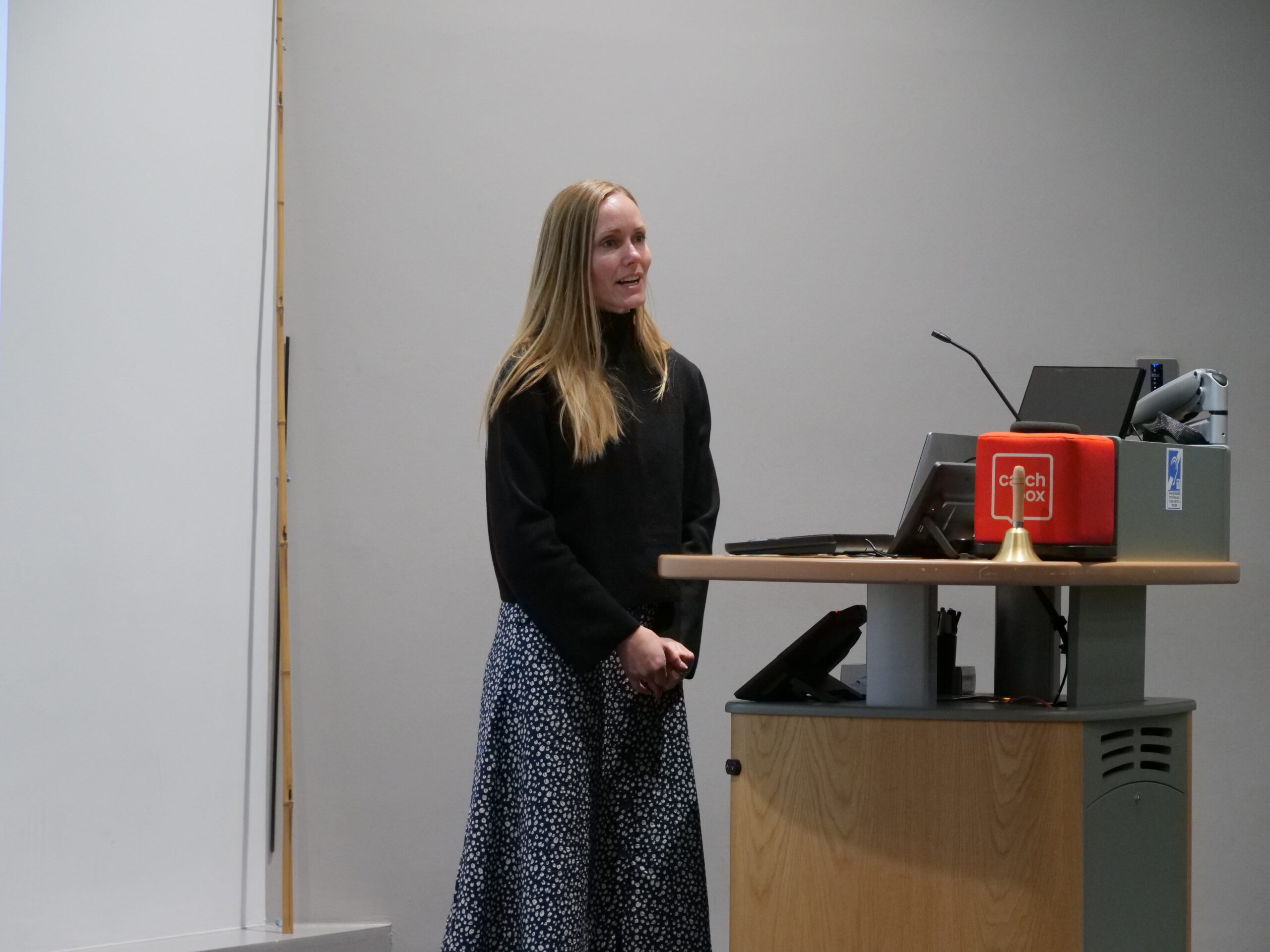A lecture room scene showing a person standing beside a lectern while speaking. The lectern holds a computer monitor, microphone, a small red box labelled ‘catch box,’ and a brass bell. The background is a plain light‑coloured wall with the edge of a projection screen visible on the left, and the room is evenly lit by ceiling lights.