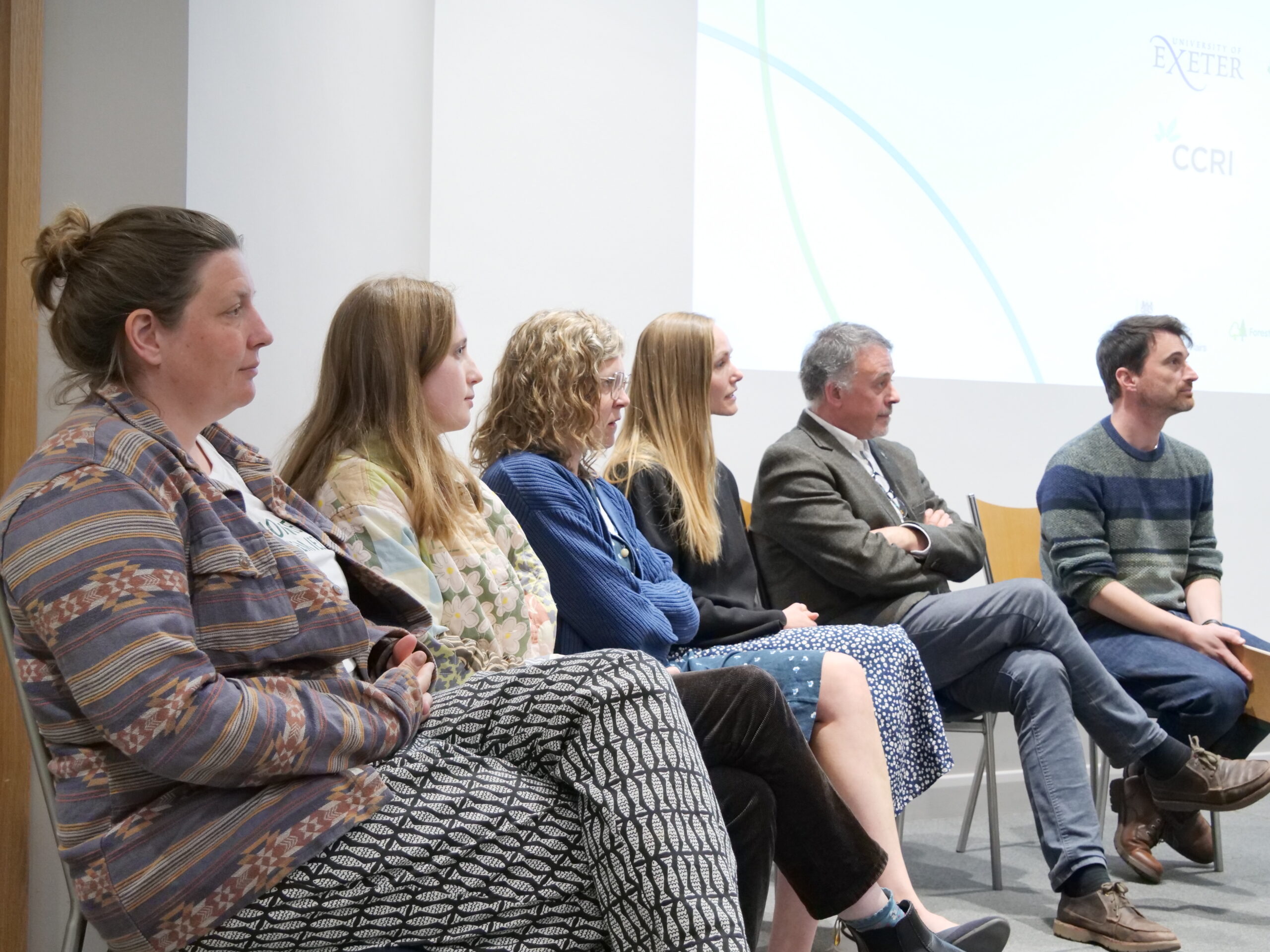 Several adults sit side‑by‑side on chairs in a lecture room during a panel discussion, viewed in profile. The individuals face toward the right, listening attentively to an off‑camera speaker. A large projected slide is partially visible on the wall behind them, showing faint logos and curved graphic lines. The room has white walls and even indoor lighting, creating a formal question‑and‑answer or discussion setting.