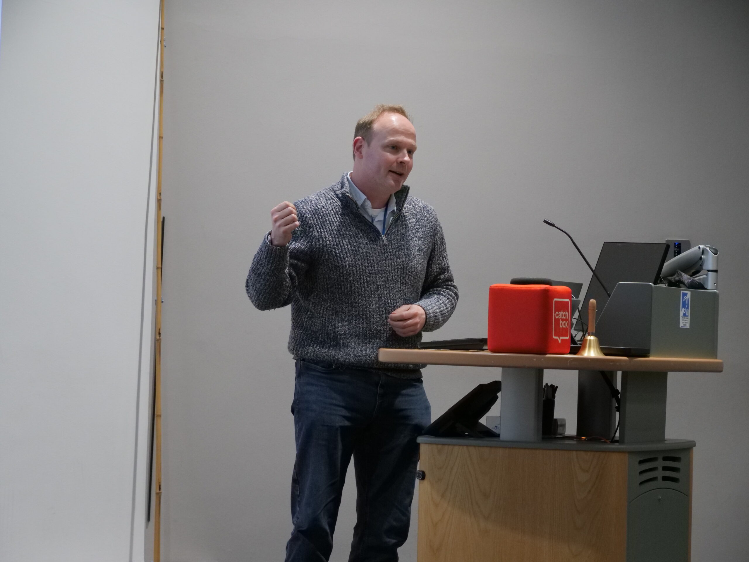 A person stands at a lectern in a lecture room, speaking while making a hand gesture. The lectern holds a computer monitor, microphone, and a small red box with the text ‘catch box,’ alongside a brass bell. A plain light‑coloured wall forms the background, and part of a projection screen is visible to the left under ceiling lighting.