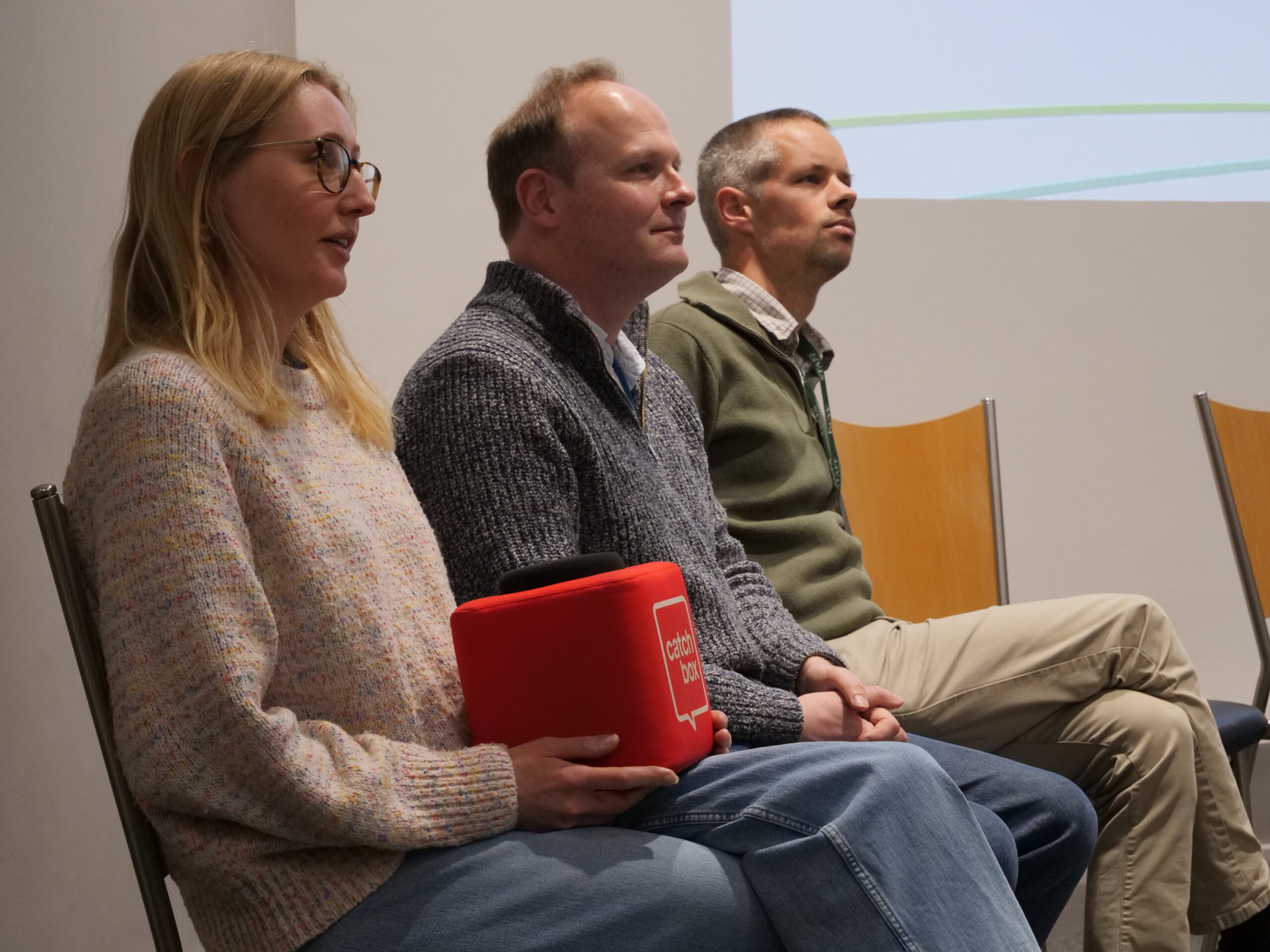 A panel discussion in a lecture room, showing three seated adults in the foreground viewed from the side. One person holds a red cube microphone labelled ‘Catch box’ while the others sit with hands folded, facing toward an off‑camera speaker or audience. The background includes a white wall with part of a projected slide showing faint curved green lines, and simple wooden chairs arranged in a row under even indoor lighting.