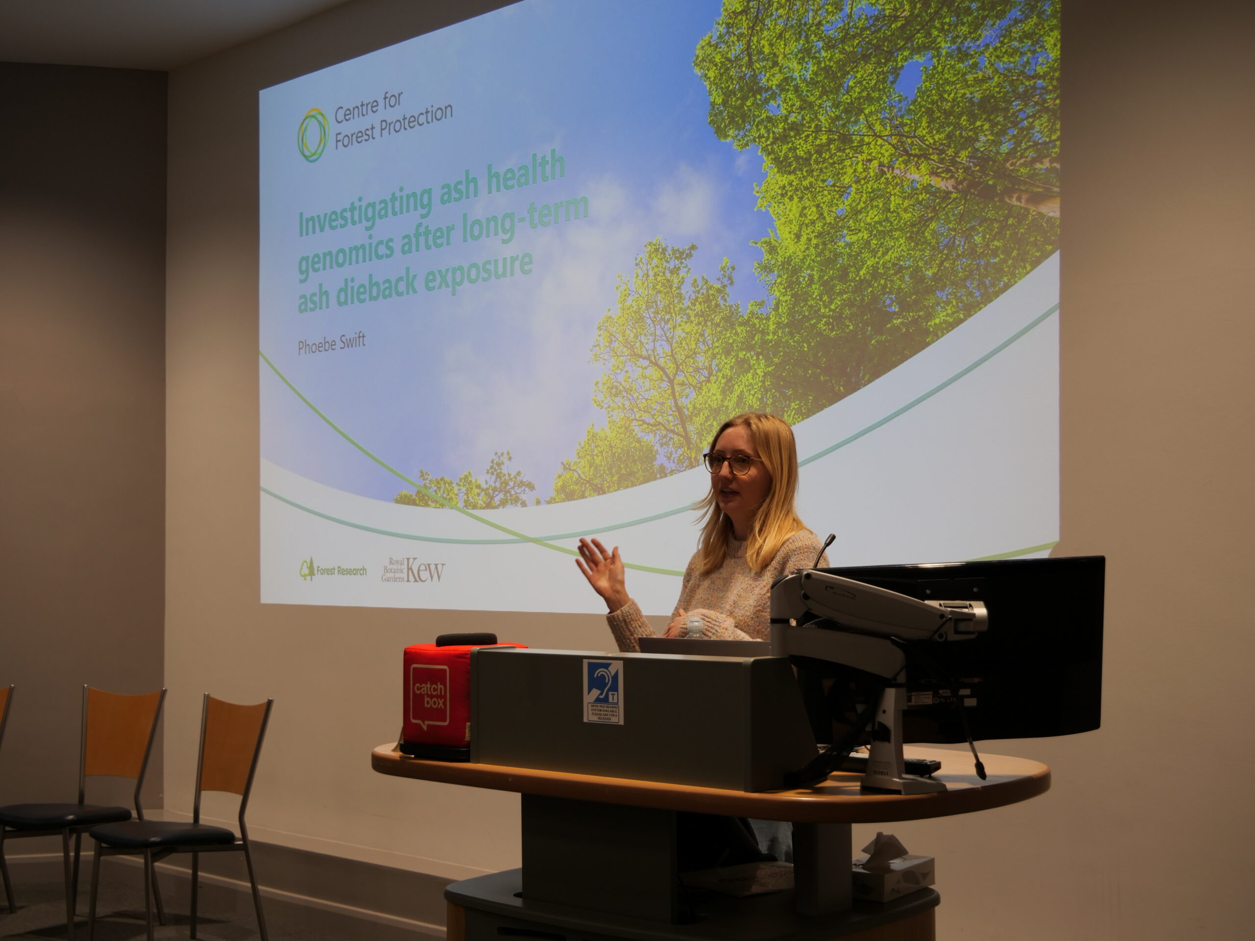 A lecture room scene showing a person standing beside a lectern while speaking. The lectern holds a computer monitor, microphone, a small red box labelled ‘catch box,’ and a brass bell. The background is a plain light‑coloured wall with the edge of a projection screen visible on the left, and the room is evenly lit by ceiling lights.