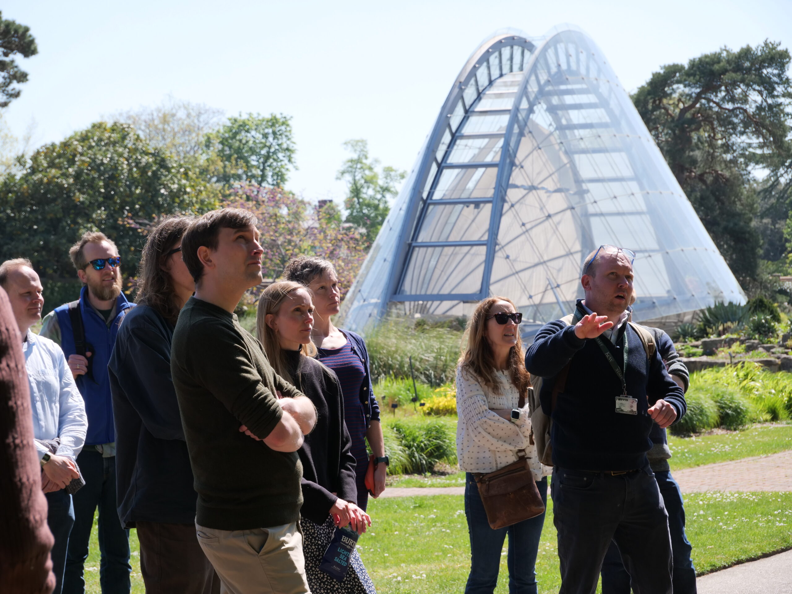 A group of adults stands outdoors on a grassy path during a guided tour, listening while one person gestures and points ahead. Behind the group is a large, curved glasshouse structure with a metal frame, set among landscaped greenery with trees, shrubs, and planted beds. The scene is brightly lit by daylight under a clear sky, with the group clustered in the foreground and the glass building rising prominently in the background.