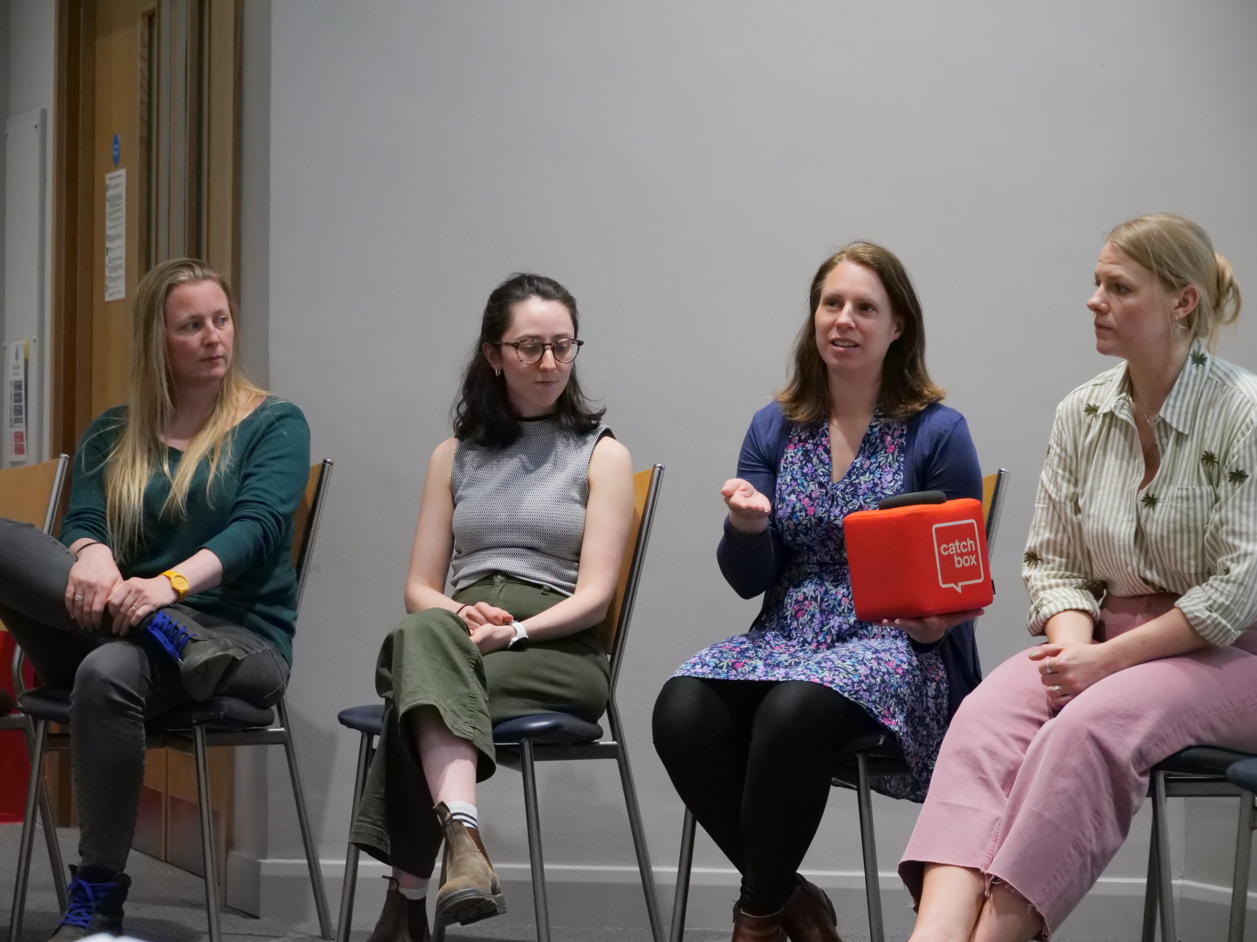 A panel discussion in a lecture room showing four adults seated in a row on simple chairs against a plain light‑coloured wall. One panellist holds a red cube microphone labelled ‘Catch box’ while speaking, and the others sit attentively facing toward the right. The setting is well lit, with a neutral background and minimal furnishings, indicating a formal question‑and‑answer session.