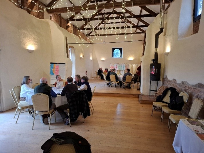 A group of people sit at round tables inside a long, rustic hall with white plaster walls, exposed wooden beams, and wooden floors. String lights hang from the ceiling, and a wood-burning stove stands along the right wall. Flip charts and papers are visible at the far end of the room, suggesting a workshop or group discussion setting.