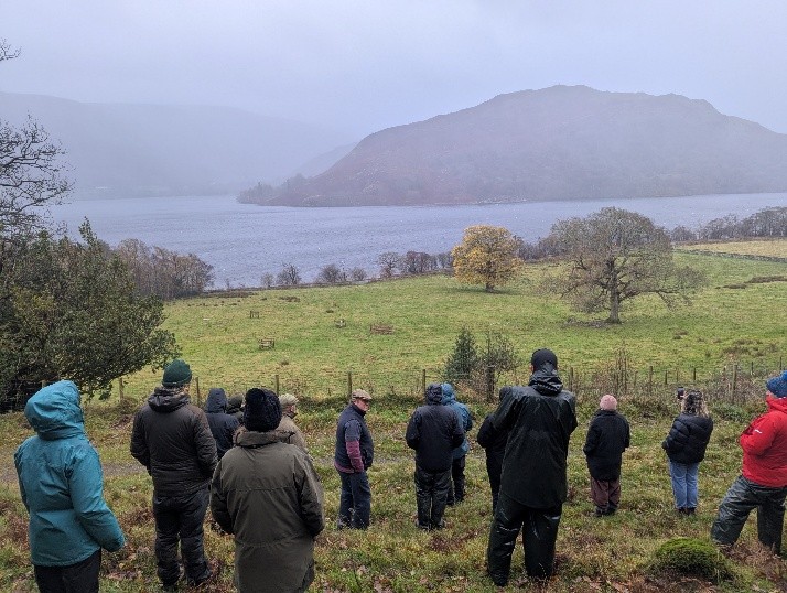A group of people wearing coats and hats stand on a grassy hillside, facing a landscape of scattered young and mature trees. Beyond the field, a wide lake stretches across the middle ground, with low, misty hills in the background under an overcast sky.