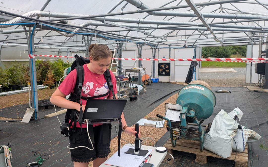 Person wearing a backpack-mounted device uses a laptop in a large greenhouse structure. Monitoring equipment and sensors are set up on a table in the foreground, with irrigation or ventilation equipment, bags, and workstations visible around the greenhouse. Red-and-white tape marks off sections of the growing area.