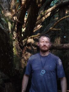 Person standing beneath a large, multi-stemmed tree with twisting, moss-covered trunks and branches. The individual is wearing a short-sleeved shirt and a round pendant on a cord around the neck. Dappled sunlight filters through the canopy, illuminating parts of the tree’s textured bark and creating strong contrasts between light and shadow in the woodland setting.